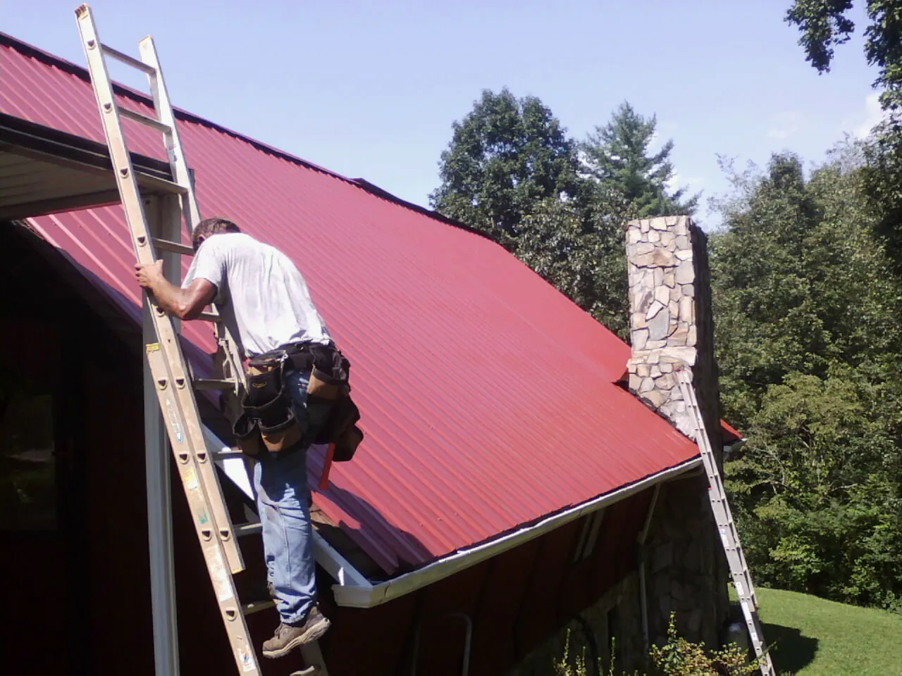 Roofer on a ladder beside a red metal roof and stone chimney — Soffit & Fascia Repair by Roofing Brothers in Carol Stream